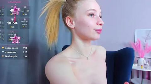 Media: Video of a fair-skinned, slender, blonde-haired woman with a high ponytail, wearing a strapless top, sitting indoors. Background includes a pink feather decoration, a digital overlay with social media stats, and a blue cushion.