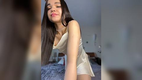 Media: Video of a young Asian woman with long, straight black hair, wearing a sheer, cream-colored camisole, leaning forward in a bedroom with a bed and a gray blanket in the background.