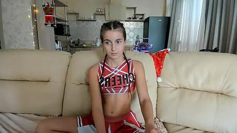 Media: Video of a young woman with fair skin and dark hair in a red and white cheerleader outfit, sitting on a beige couch in a modern living room.