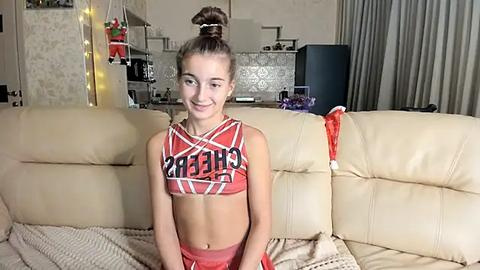 Media: Video of a smiling young girl with light skin, brown hair in a high ponytail, wearing a red and white \"Cheer\" crop top and matching shorts, sitting on a beige leather sofa in a cozy living room.