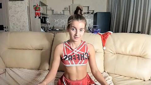 Media: Video of a young, athletic Caucasian woman with light skin, brown hair in a high ponytail, wearing a red and white \"Chargers\" cheerleading crop top and shorts, sitting on a beige leather couch in a cozy living room with Christmas decorations.