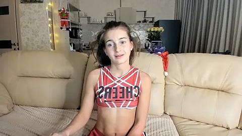 Media: Video of a young Caucasian woman with fair skin, light brown hair, wearing a red and white cheerleading crop top with \"CHEER\" on it, sitting on a beige leather couch in a modern living room.