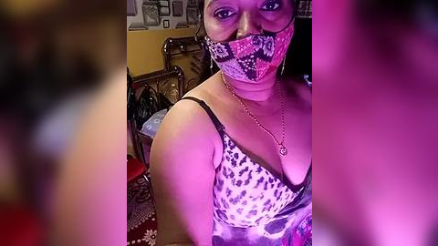 Media: Video of a woman with medium brown skin, wearing a leopard print face mask and black bra, sitting in a dimly-lit room with a bed, and a blurred background.