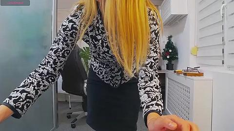 Media: Video of a blonde woman with long hair, wearing a black and white floral blouse and a navy skirt, reaching out in a modern office with white walls and furniture.