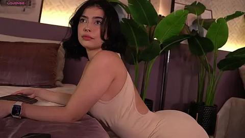 Media: Video of a young woman with medium skin tone, dark hair, and brown eyes, lying on a bed, wearing a beige, backless dress. The room features a lavender headboard, green plants, and a warm, ambient lighting.