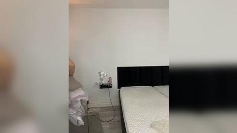 Media: A video of a minimalist, modern bedroom with a single bed, white bedding, and a small black bedside table with a lamp. The walls are white, and the floor is light gray.