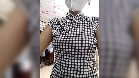 Media: Video of a person wearing a black-and-white checkered dress with short sleeves, a white mask covering their face, and a white headband. The background features a blurred indoor setting with a ceiling and furniture.