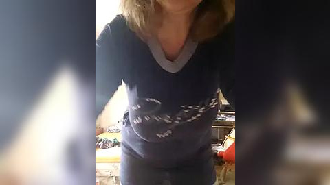 Media: Video of a light-skinned woman with shoulder-length blonde hair, wearing a dark blue V-neck sweater with white text, standing indoors. Background features a cluttered room with a bed and various objects.