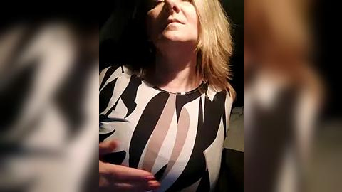 Media: Video of a woman with shoulder-length blonde hair, wearing a black and white striped blouse, sitting in a dimly lit room, with a blurred figure in the foreground.