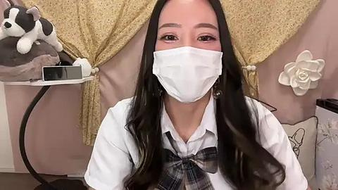 Media: Video of a young Asian woman with long black hair, wearing a white face mask, white shirt, and plaid tie, in a cozy, girly room with pastel colors and stuffed animal decor.