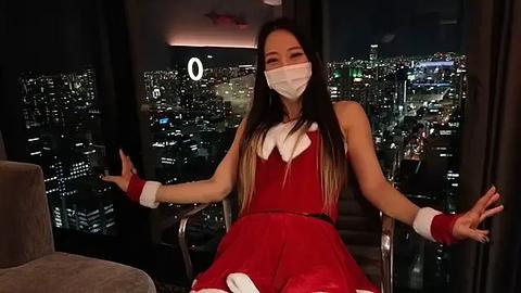 Media: Video of an Asian woman in a red Santa dress with white fur trim, white mask, and red cuffs, sitting in a modern, dark-lit room with a city skyline view.