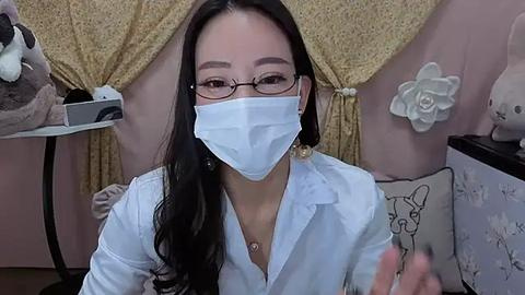 Media: A video of an Asian woman with long black hair, wearing a white mask, glasses, and a white shirt, seated in a cozy room with yellow curtains, stuffed animals, and floral decor.