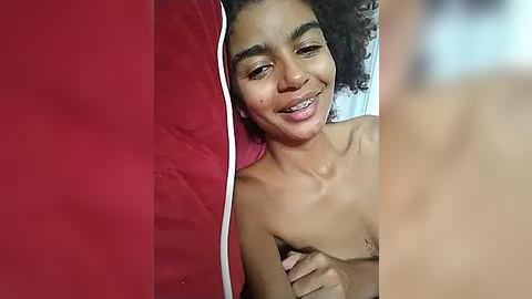 Media: Video of a smiling, topless Black woman with curly hair, lying on a red pillow against a red wall, with a blurry, light-skinned man's arm in the foreground.