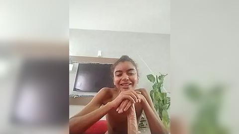 Media: Video of a smiling, topless young woman with light brown skin, dark hair in a ponytail, hands clasped, in a modern, minimalist room with a flat-screen TV, potted plant, and blurred foreground.