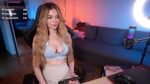 Media: Video of a young woman with long, wavy blonde hair, wearing a low-cut light blue crop top and high-waisted white shorts, sitting in a gaming chair. Background features a gray couch, a ring light, and a laptop.