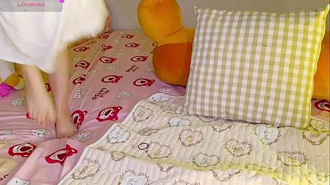 Media: Video of a toddler in a white robe, barefoot, standing on a bed with a pink quilt adorned with red heart patterns. Surrounding the bed are plush toys and a yellow checkered pillow.