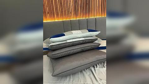 Media: Video of a neatly made bed with a gray headboard, featuring three pillows in varying shades of gray and blue, set against a warm, wooden wall with vertical slats.