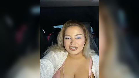 Media: Video of a smiling, light-skinned woman with blonde hair, wearing a pink bra, white cardigan, and purple eyeshadow, sitting in a car with dark surroundings.