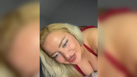 Media: A video of a blonde woman with fair skin and medium-sized breasts, wearing red lingerie, smiling seductively. The background is blurred, focusing on her face and chest.