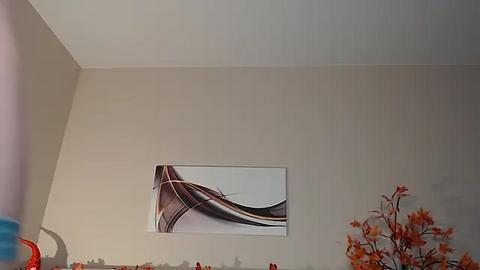 Media: A video of a minimalist room with beige walls featuring a modern abstract painting of swirling, wavy lines in black, white, and gray. The right side displays a vase of orange and red flowers.