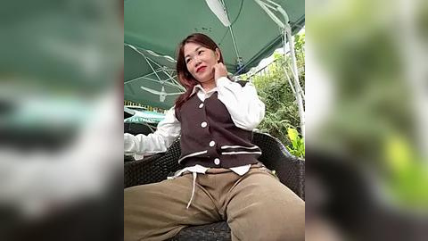 Media: Video of an Asian woman with long brown hair, wearing a white shirt, brown vest, and beige pants, sitting outdoors under a green umbrella, smiling.