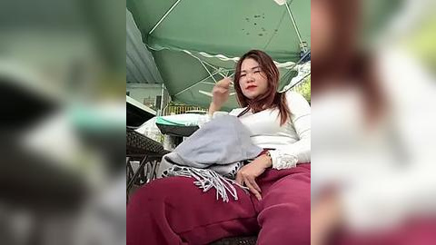 Media: A video of an Asian woman with long brown hair, wearing a white sweater and maroon pants, sitting under a green umbrella at an outdoor caf\u00e9, with blurred background details.