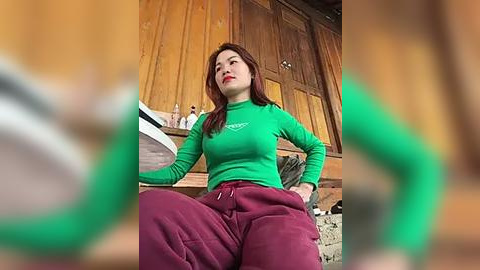 Media: Video of a young woman with long brown hair, wearing a green sweater and maroon pants, sitting on a wooden bench inside a rustic cabin with wooden walls and a sink.