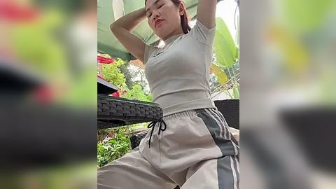 Media: Video of an East Asian woman in casual beige and gray athletic wear, standing on a black wicker chair, arms raised, in a lush, green garden with a blurred background.