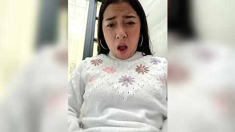 Media: Video of a young Asian woman with long black hair, wearing a white sweater with floral embroidery, looking shocked, indoors near a window.