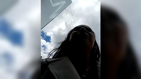 Media: Video of a woman with long, dark hair, wearing a dark jacket, leaning back, head tilted, and eyes closed, under a cloudy sky with a blue, white, and gray gradient.