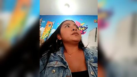 Media: Video of a Latina woman with long dark hair, wearing a denim jacket, looking upward in a brightly lit room with vibrant, colorful murals on the walls.