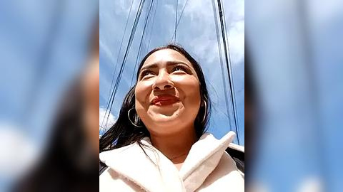 Media: Video of a smiling, light-skinned woman with straight black hair, wearing a white hoodie, against a clear blue sky with power lines in the background.