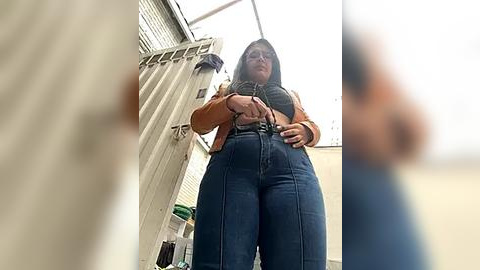 Media: Video of a tall, plus-sized woman with long dark hair, wearing a brown jacket over a plaid shirt, high-waisted jeans, and glasses, standing in a garage with a white door and window.