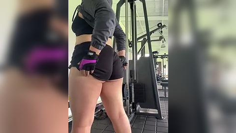 Media: Video of a woman in a gym, wearing black sports bra, black shorts, and black gloves. She is adjusting her shorts while standing in front of gym equipment.