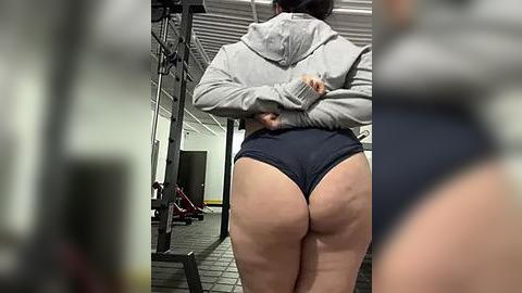 Media: Video of a woman in a gym, lifting weights. She has a curvy figure, wearing a gray hoodie and black high-cut workout shorts, showcasing her round buttocks. The gym is dimly lit with equipment in the background.