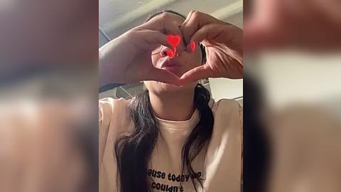 Media: Video of a young woman with long dark hair, wearing a white T-shirt with \"House of Cards\" printed on it. She forms a heart shape with her hands over her face, with red heart emojis replacing her eyes. Background is blurred, indoor setting.