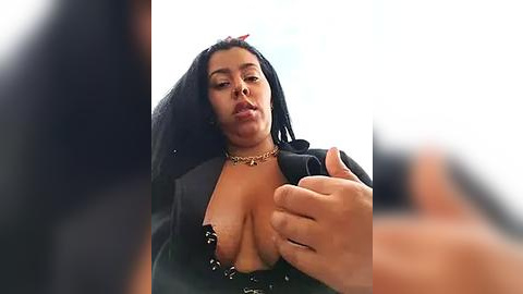 Media: A video of a curvaceous, dark-skinned woman with large breasts, wearing a black top with a plunging neckline, and a gold choker. She is in a suggestive pose, with her hand placed on her breast.