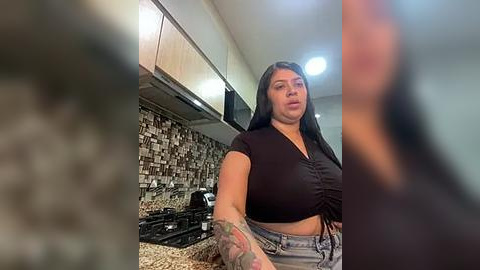 Media: Video of a plus-sized woman with long black hair, wearing a black crop top and high-waisted jeans, standing in a modern kitchen with a stone backsplash and black appliances.