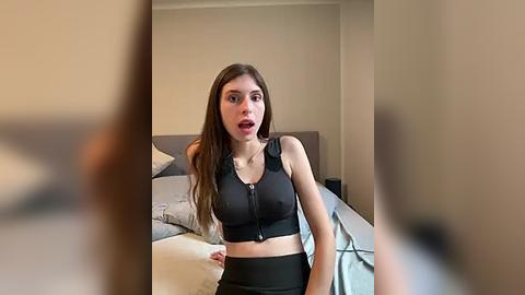 Media: Video of a young woman with long brown hair, fair skin, wearing a black sports bra and black shorts, surprised expression, in a bedroom with beige walls and a gray bed.