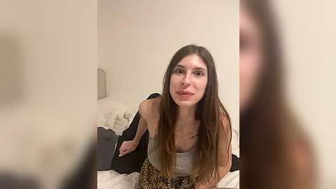 Media: A video shows a young woman with long brown hair, wearing a leopard-print dress, sitting on a bed with white sheets in a dimly lit bedroom.