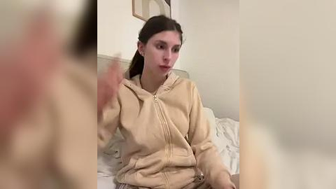 Media: A video of a young woman with long brown hair, wearing a beige hoodie, sitting on a bed with white sheets, gesturing with her left hand. The background features a neutral-colored wall with a framed picture.