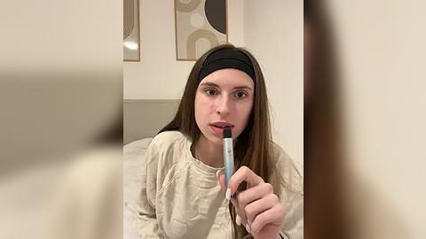 Media: Video of a young woman with long brown hair, wearing a beige sweater and a black headband, applying lipstick with a light blue tube, in a modern bedroom with beige walls and abstract art.