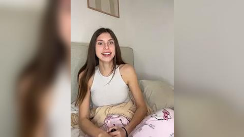 Media: A video of a young woman with long brown hair, wearing a white tank top and pink pajama pants, sitting on a bed in a minimalist room.