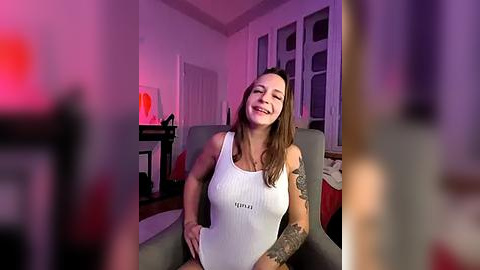 Media: Video of a smiling, tattooed woman in a white tank top, sitting in a grey armchair, in a dimly lit, modern living room with purple lighting.