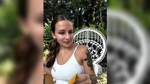 Media: Video of a young woman with light skin, brown hair in a braid, wearing a white tank top, holding a banana, seated on a white, ornate outdoor chair in a lush, green garden.