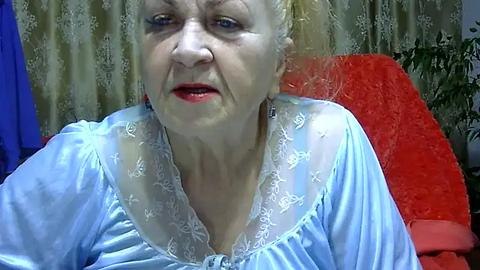 Media: Video of an elderly white woman with fair skin, wearing a sheer, light blue nightgown with lace detailing. She has short, blonde hair, red lipstick, and sits in a red chair with a beige curtain background.