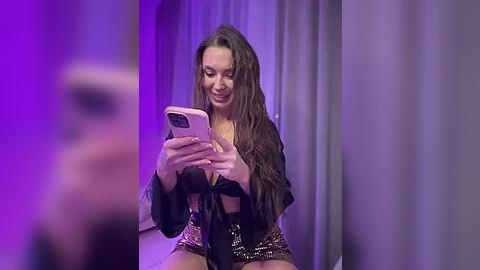 Media: Video of a young woman with long, wavy brown hair, wearing a black tie-front top and sequined shorts, smiling at her smartphone, in a purple-lit room with gray curtains.