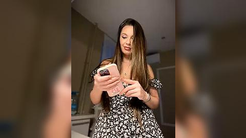 Media: Video of a smiling, light-skinned woman with long, straight brown hair, wearing a black and white floral dress, holding a smartphone, in a modern, dimly lit room.