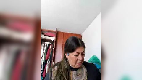 Media: Video of a woman with dark hair and glasses, wearing a black sweater and patterned scarf, standing in a cluttered closet with hanging clothes and wooden shelves.