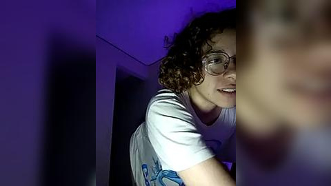 Media: Video of a young person with curly hair and glasses, wearing a white T-shirt, peering through a narrow gap in a dark room illuminated by a purple light.